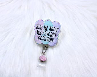 May include: A badge reel with a pastel cloud-shaped design. The badge reel has the text "ASK ME ABOUT MY FAVORITE POSITIONS" in black. The badge reel has a glittery pink accent and a clear plastic holder.