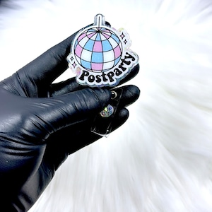 May include: A badge reel featuring a circular acrylic topper with a glittery finish. The design displays a disco ball with light blue and pink panels, accompanied by the text Postparty in a bold black font and small star icons on the sides.