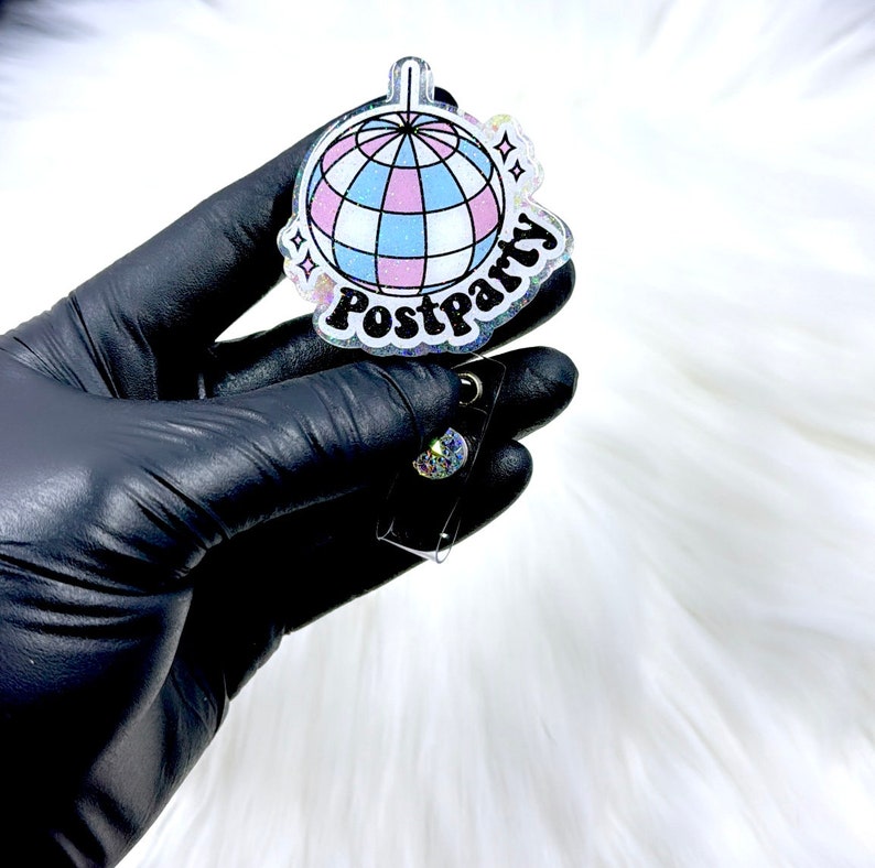 May include: A badge reel featuring a circular acrylic topper with a glittery finish. The design displays a disco ball with light blue and pink panels, accompanied by the text Postparty in a bold black font and small star icons on the sides.