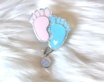 Puede incluir: Un portagafete de plástico transparente con un dije de pie de bebé de purpurina rosa y azul con un pequeño corazón blanco.