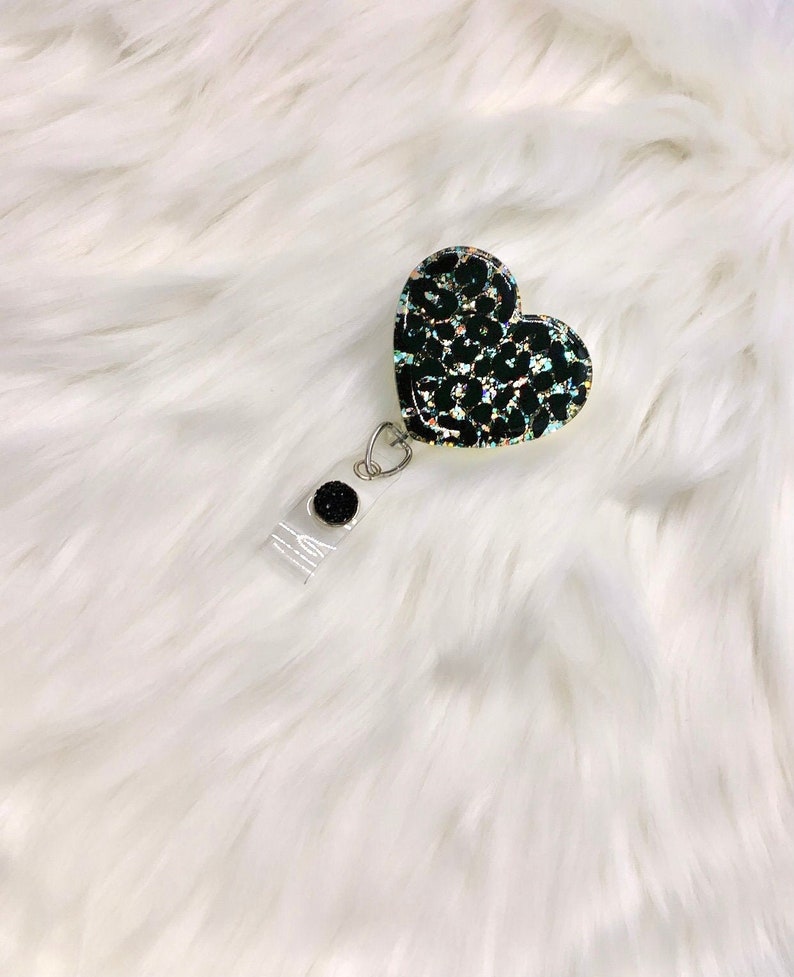 Puede incluir: Un portagafete en forma de coraz&oacute;n negro y dorado con un dise&ntilde;o de estampado de leopardo. El portagafete tiene un carrete retr&aacute;ctil de pl&aacute;stico transparente y un bot&oacute;n negro.