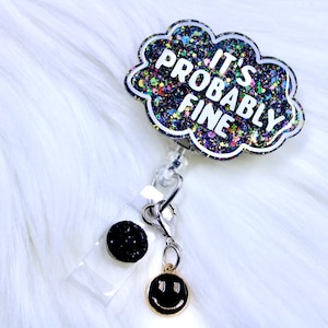 Pu&ograve; includere: Un portabadge con un design a forma di nuvola che recita "IT'S PROBABLY FINE" in lettere bianche. La nuvola &egrave; piena di glitter colorati. Ha un bottone nero glitterato e un ciondolo a forma di faccina sorridente dorata.