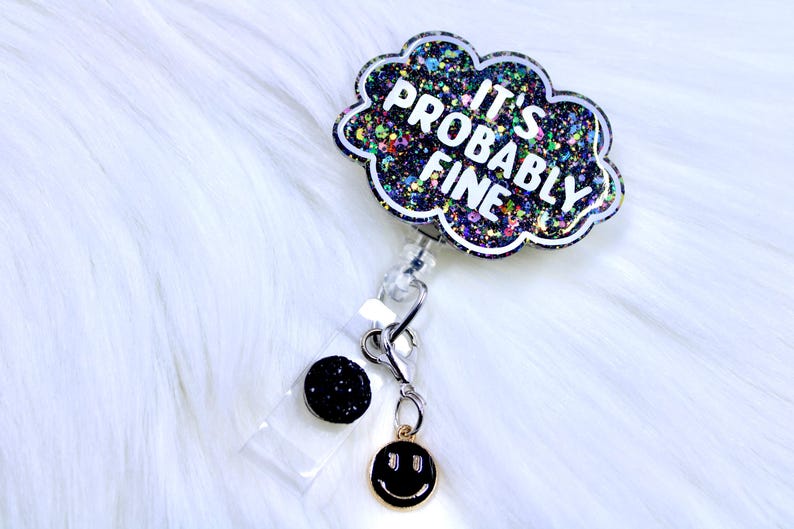 Pu&ograve; includere: Un portabadge con un design a forma di nuvola che recita "IT'S PROBABLY FINE" in lettere bianche. La nuvola &egrave; piena di glitter colorati. Ha un bottone nero glitterato e un ciondolo a forma di faccina sorridente dorata.