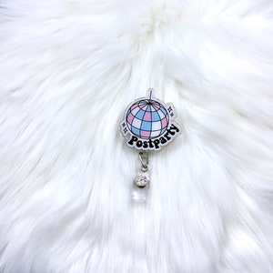 May include: A badge reel featuring a circular acrylic topper with a disco ball graphic in shades of pink, blue, and white. The word Postparty is printed in black script below the disco ball. A clear plastic strap with a small rhinestone is attached.