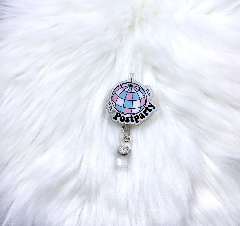 May include: A badge reel featuring a circular acrylic topper with a disco ball graphic in shades of pink, blue, and white. The word Postparty is printed in black script below the disco ball. A clear plastic strap with a small rhinestone is attached.