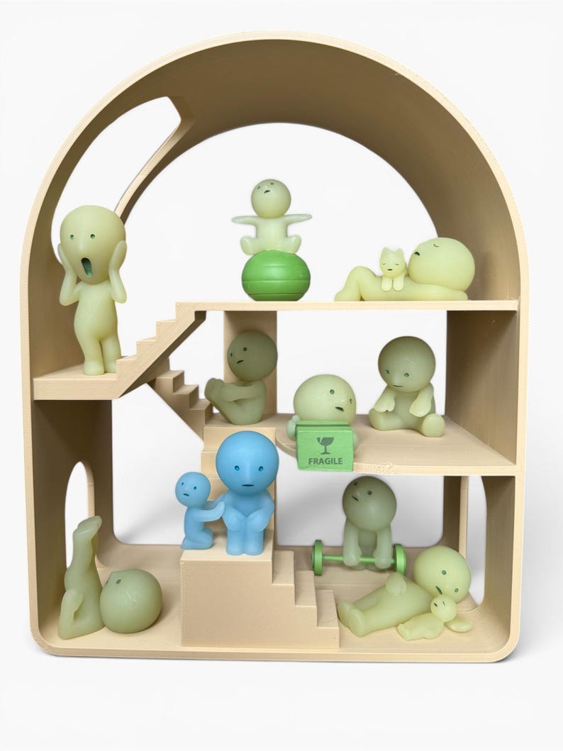 May include: A beige wooden dollhouse with multiple levels, housing various small, stylized figurines in shades of green and blue. Some figurines are posed in different activities, such as lifting weights or reclining. A green box with the word "FRAGILE" is also present.