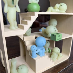 May include: A miniature dollhouse with multiple levels, featuring various small, stylized figures in shades of green and blue. Some figures are posed on stairs, while others interact with each other. A small green box with the word "FRAGILE" is also present.