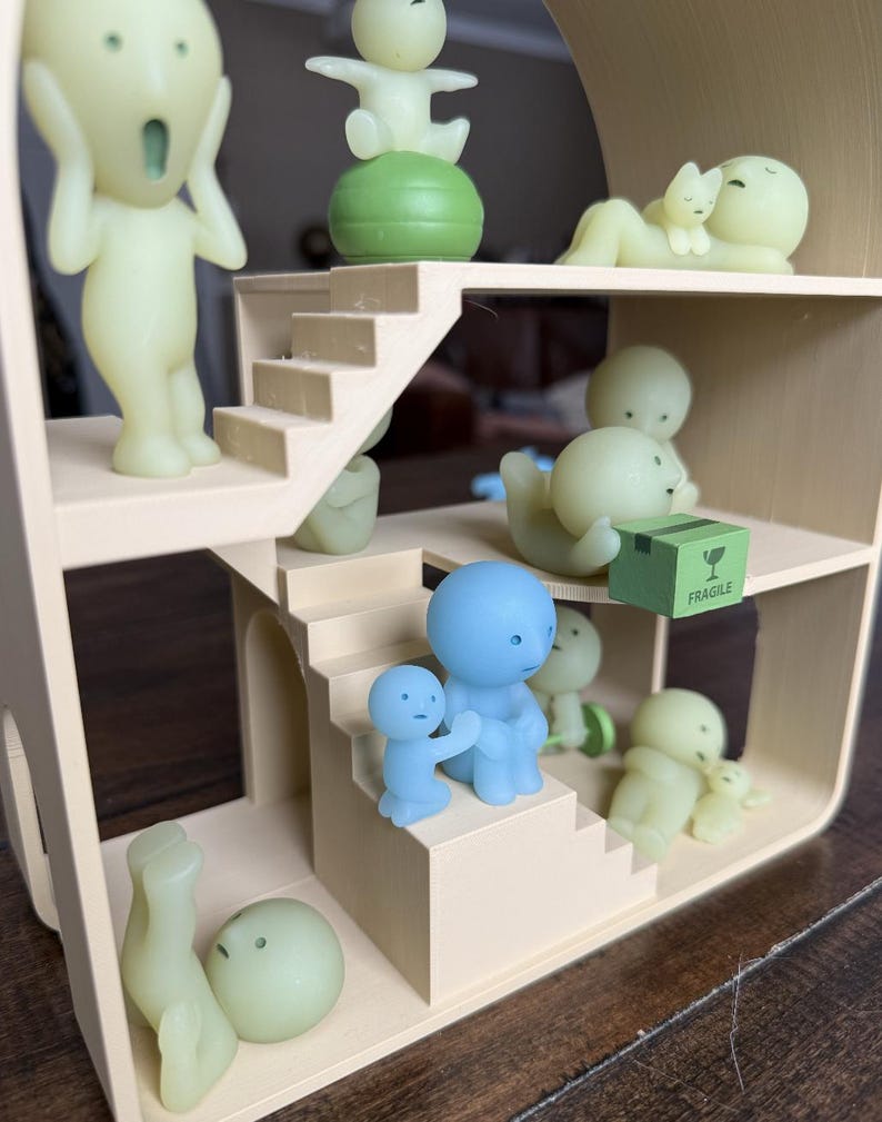 May include: A miniature dollhouse with multiple levels, featuring various small, stylized figures in shades of green and blue. Some figures are posed on stairs, while others interact with each other. A small green box with the word "FRAGILE" is also present.
