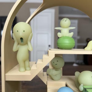 May include: A whimsical, multi-level structure with various small, light green figurines. One figurine is posed with hands on its face, another is seated, and others are standing. The structure is beige and has stairs and rounded architectural elements.