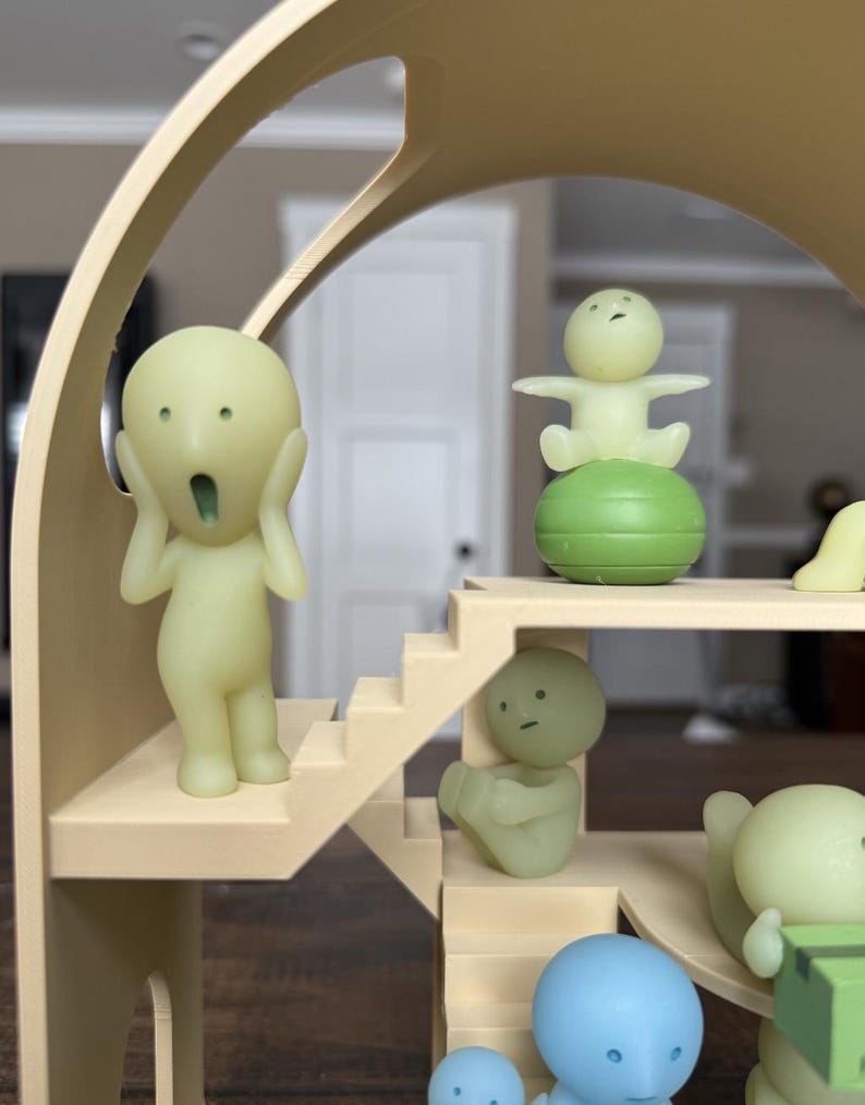 May include: A whimsical, multi-level structure with various small, light green figurines. One figurine is posed with hands on its face, another is seated, and others are standing. The structure is beige and has stairs and rounded architectural elements.
