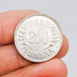 May include: A silver Finnish coin with a value of 200 markkaa. The coin features a design of stylized pine trees and the text "200 MARKKAA".