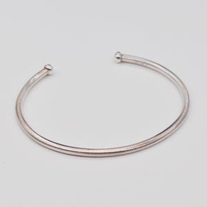 Könnte beinhalten: Ein silberfarbenes Armreif-Armband mit einem strukturierten, geriffelten Design. Das offene Armband hat an jedem Ende eine kleine, abgerundete Kugel. Der Schmuck wird vor einem schlichten, hellfarbigen Hintergrund gezeigt.