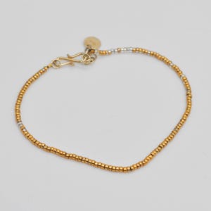 Peut inclure: Un bracelet délicat en perles dorées avec un fermoir à crochet et un petit charme circulaire. Le bracelet présente un mélange de perles dorées et argentées. Le bijou est conçu pour un usage quotidien.