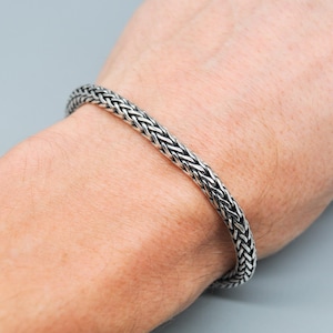 Könnte beinhalten: Ein silberfarbenes geflochtenes Armband. Das Armband besteht aus ineinandergreifenden Metallgliedern, die ein strukturiertes, gewebtes Aussehen erzeugen. Das Armband wird am Handgelenk getragen und zeigt sein Design und seine Handwerkskunst.