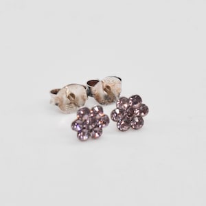 May include: A pair of stud earrings featuring a flower-shaped design composed of small, light purple circular crystals. The earrings include metallic silver-toned backings with visible markings, positioned behind the crystal flower fronts.