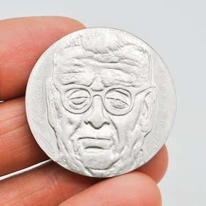 May include: Silver coin with a portrait of a man wearing glasses. The coin has the dates 1810-1412-1930 inscribed around the edge.