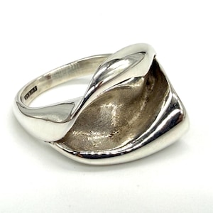 May include: A polished silver ring featuring a wide, sculptural band with a smooth, reflective outer surface and a textured, concave interior section. The metal band curves into a twisted, asymmetrical shape that wraps around the finger.