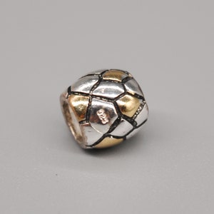 May include: A silver and gold bead with a geometric design. The bead is cube-shaped, featuring silver and gold panels. The number "925" is stamped on the side. The bead is set against a grey backdrop. The bead is likely a charm or pendant.