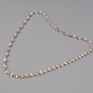 Puede incluir: Un collar plateado con un diseño de espiral repetitivo. El collar tiene una cadena delicada y un cierre. El collar se muestra sobre un fondo gris.