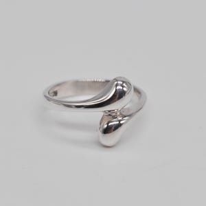 May include: A polished silver-toned ring featuring a bypass design with two smooth, rounded, teardrop-shaped ends that overlap slightly. The metal surface is highly reflective, creating a sleek, continuous appearance across the band and terminals.