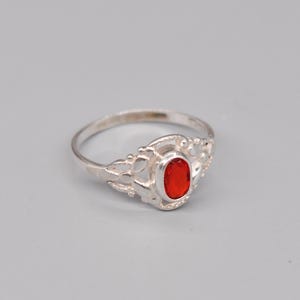 May include: A silver ring featuring a vibrant red, oval gemstone. The ring has an ornate, openwork design surrounding the stone. The band is slender and polished. The ring is set against a pale grey backdrop.
