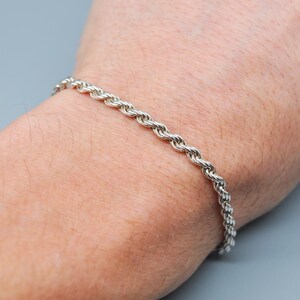 Könnte beinhalten: Ein silbernes Armband mit Seilkette. Das Armband besteht aus eng gewebten Gliedern, die eine strukturierte Oberfläche bilden. Es wird an einem Handgelenk getragen, um sein Design hervorzuheben. Der Hintergrund ist einfarbig hellgrau.