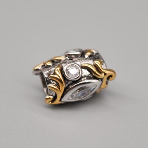 May include: A silver and gold charm with a detailed design featuring a swirl pattern and clear cubic zirconia stones.