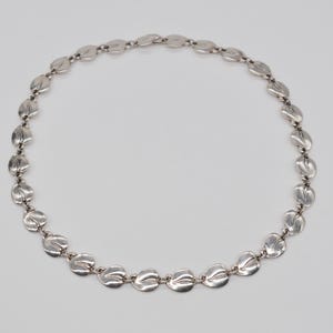 May include: A silver necklace with a repeating pattern of leaf-shaped links. The links are connected by small rings, forming a continuous chain. The necklace is displayed against a plain, light background.