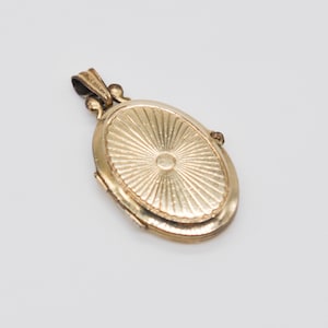 May include: A gold-colored oval locket pendant with a sunburst design on the front. The locket has a small loop at the top for a chain and a hinged opening. The pendant is likely made of metal and has a vintage or antique appearance.