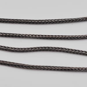 Puede incluir: Un collar de cadena de cola de zorro metálico de color gris oscuro con un patrón de eslabones trenzados y tejidos apretadamente. La cadena tiene un perfil redondeado y suave con una textura consistente en toda su longitud, mostrando un brillo sutil.