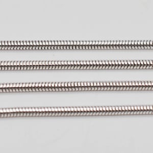 Könnte beinhalten: Vier parallele, silberfarbene Schlangenketten, die horizontal angeordnet sind. Jede Kette weist ein glattes, flexibles und abgerundetes Design mit ineinandergreifenden Gliedern und einer polierten Oberfläche auf.