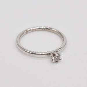 May include: A silver ring featuring a single, round, clear gemstone. The thin, smooth ring band has a small inscription. The gemstone is held in a four-prong setting. The ring is shown on a white surface.