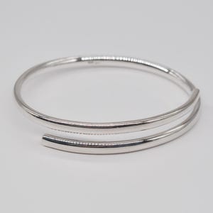 Puede incluir: Un brazalete plateado con un diseño de doble banda. El brazalete tiene un acabado liso y pulido y una estética minimalista. Las bandas se curvan, creando un aspecto elegante y moderno. La joya está colocada sobre un fondo blanco liso.