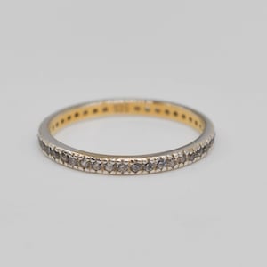 Puede incluir: Un anillo fino y circular que presenta una hilera continua de pequeñas piedras redondas y transparentes engastadas en un exterior de metal plateado. El interior de la banda es de tono dorado y lleva el sello 925, indicando plata de ley.