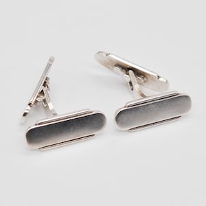 May include: A pair of silver cufflinks with a rectangular design. The cufflinks have a polished finish and a hinged back for secure fastening. The design is simple and elegant, suitable for formal wear.