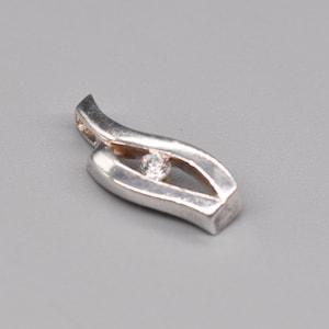 May include: A silver pendant with a flowing, leaf-like design. The pendant features a small, round, clear gemstone set in the center. The metal has a polished finish, and the overall shape is elongated and curved.