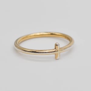 May include: A gold and silver ring with a cross design. The ring has a thin band with a gold finish and a silver cross accent. The cross is positioned on the front of the ring, creating a simple and elegant appearance.