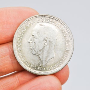 Puede incluir: Una moneda de plata con el perfil de un hombre, rodeada de texto. La moneda lleva la inscripción "GUSTAF V SVERIGES KONUNG" y otras palabras. La moneda se sostiene entre los dedos, mostrando sus detalles.