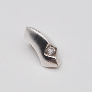 May include: A curved, silver-toned ring with a single, clear gemstone set in a star-shaped bezel. The ring has a smooth, polished finish and a modern design. The gemstone is round and sparkles.