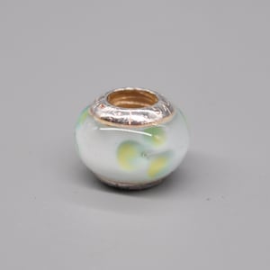 May include: A white glass bead with green and yellow swirls. The bead has a silver and gold metal band around the center.