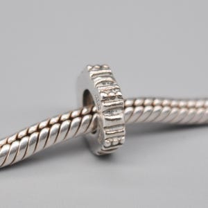 Op de afbeelding: Een zilveren bedel met een getextureerd, geometrisch ontwerp. De bedel is cilindrisch met een centraal gat en is geregen op een zilveren ketting. Het oppervlak van de bedel heeft een gedetailleerd patroon en de algehele esthetiek is modern en minimalistisch.