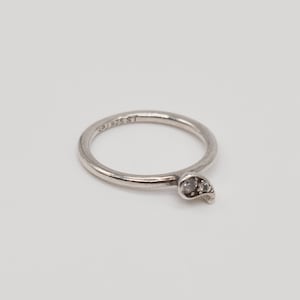 Puede incluir: Un anillo plateado con un pequeño diseño en forma de hoja. El anillo presenta dos pequeñas piedras preciosas transparentes. La banda es delgada y tiene las marcas "SP 925 ST". El anillo está sobre un fondo blanco.