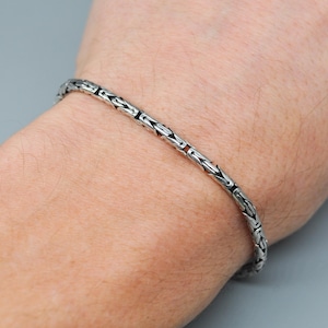 Pode incluir: Um bracelete de corrente de prata com um design de corrente de caixa. O bracelete está no pulso de uma pessoa.