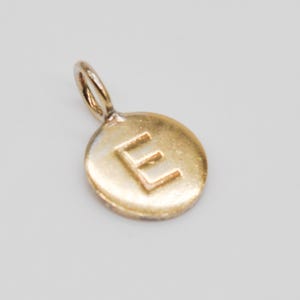May include: A gold-colored pendant with the letter "E" engraved on a circular charm. The pendant has a small loop at the top for attaching to a chain. The charm is approximately 0.5 inches in diameter.