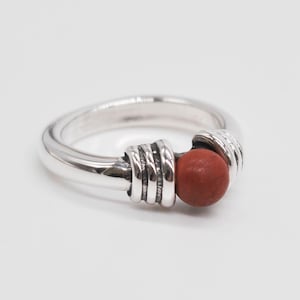 May include: A polished silver ring featuring a smooth, round, reddish-brown stone bead set between two decorative silver bands with three horizontal grooves each. The metal has a bright, reflective finish and a sturdy, rounded band design.