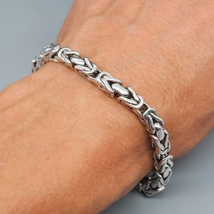 Könnte beinhalten: Ein silbernes Kettenarmband mit einem Box-Ketten-Design. Das Armband befindet sich am Handgelenk einer Person.