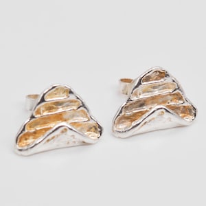 May include: A pair of silver-toned stud earrings. Each earring has a unique, organic shape with a layered design and a warm, golden-yellow interior. The earrings are set against a plain white background.