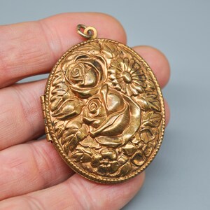Op de afbeelding: Ovaal goudkleurig medaillon met een bloemmotief. Het medaillon heeft reliëf rozen en andere bloemen, met een getextureerde rand. Het medaillon heeft een kleine lus aan de bovenkant voor een ketting.