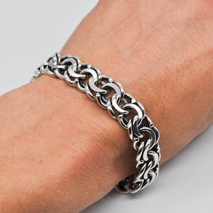Könnte beinhalten: Ein silberfarbenes Armband mit einem dicken, ineinandergreifenden Kettendesign. Das Armband wird am Handgelenk getragen und hat eine polierte, reflektierende Oberfläche. Die Glieder sind abgerundet und erzeugen ein kühnes, strukturiertes Aussehen. Der Schmuck ist ein Statement-Stück.
