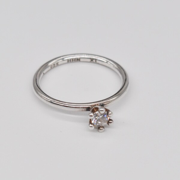 May include: A silver ring with a small, clear gemstone. The ring has a delicate band and a six-prong setting holding the round-cut stone. The ring is on a white background.